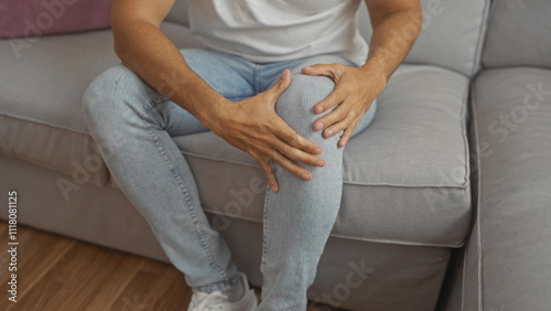 Fotografie A hispanic man sitting on a gray couch in a living room, holding his knee in apparent discomfort and wearing casual clothing