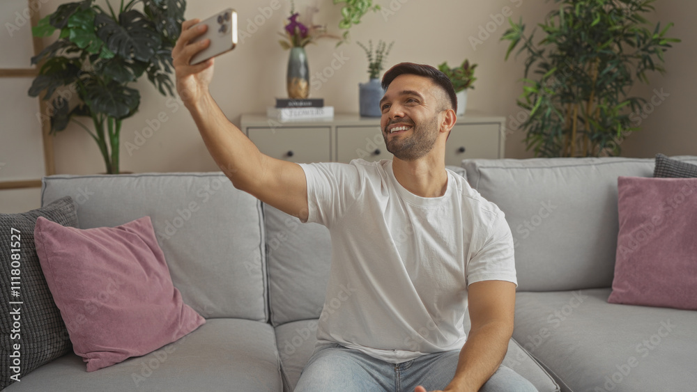 A handsome young hispanic man with a beard, taking a selfie in the living room of his apartment, sitting on a comfortable couch with lush greenery in the background. A handsome young hispanic man with a beard, taking a selfie in the living room of his apartment, sitting on a comfortable couch with lush greenery in the background.