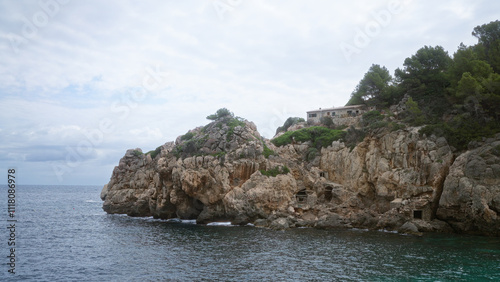 Rocky coastline with scenic cliffs and a rustic stone house surrounded by lush greenery, cala deia, mallorca, with the mediterranean sea in the foreground under a cloudy sky