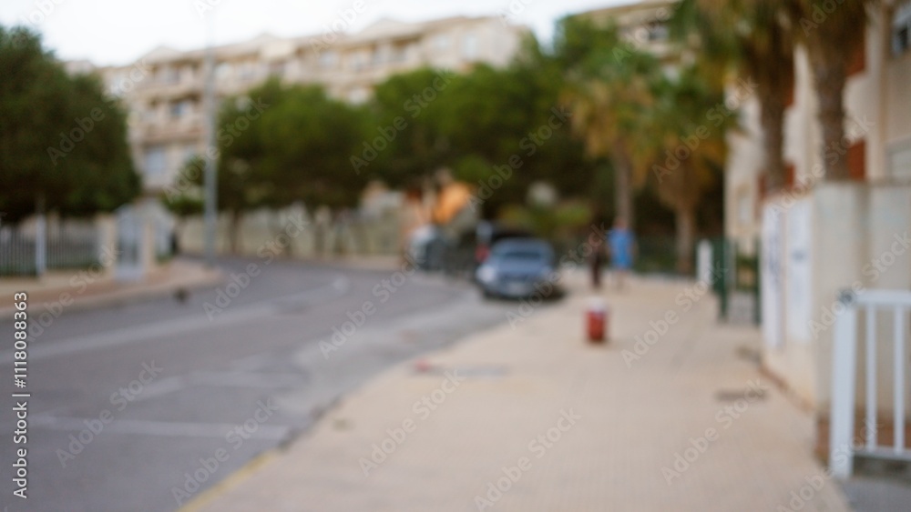 Blurry defocused outdoor scene with blurry buildings, streets, trees, and a few blurred people walking on a sidewalk in a neighborhood. Blurry defocused outdoor scene with blurry buildings, streets, trees, and a few blurred people walking on a sidewalk in a neighborhood.