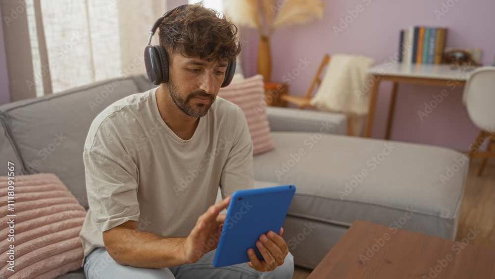 Young man using tablet while wearing headphones seated in a cozy living room, suggesting a relaxed home environment with a couch, soft pillows, and a desk in the background. Young man using tablet while wearing headphones seated in a cozy living room, suggesting a relaxed home environment with a couch, soft pillows, and a desk in the background.