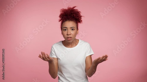 Portrait African American redhead curly woman spreads hands looks camera, say what you want from me. African female in white t-shirt on pink background with copy space. People emotions lifestyle