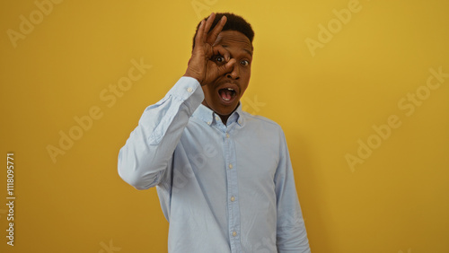 Young man makes an ok gesture in front of his eye, standing against a vibrant yellow background, expressing positivity and playfulness.