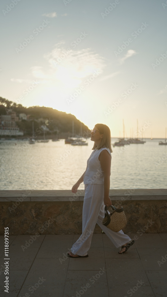 Woman walking along a scenic waterfront during sunset in mallorca, spain, holding a bag with sailboats and hilly coastline in the background