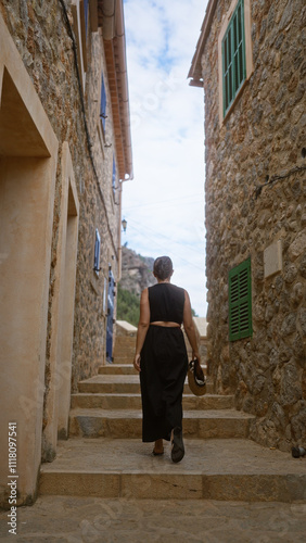 Young woman exploring narrow street in port de valldemossa, mallorca, spain, showcasing traditional stone buildings and clear blue sky above.