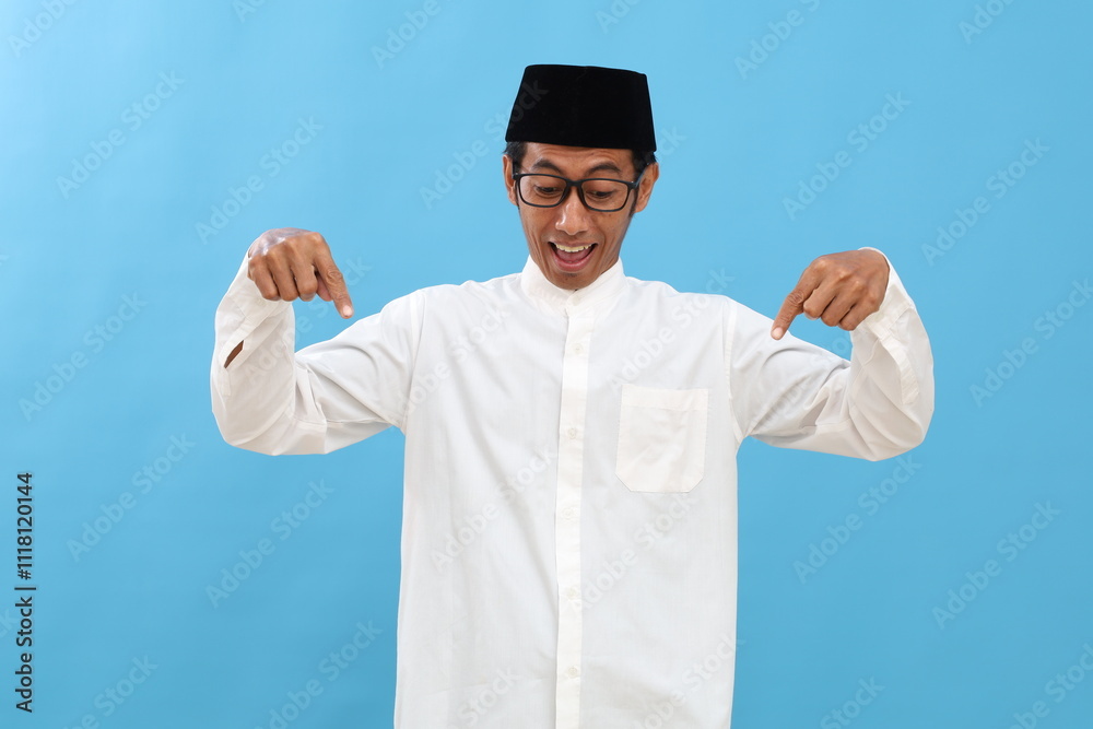 Asian muslim man feeling shocked, open-mouthed and amazed, looking and pointing downwards in disbelief and surprise against blue background Asian muslim man feeling shocked, open-mouthed and amazed, looking and pointing downwards in disbelief and surprise against blue background