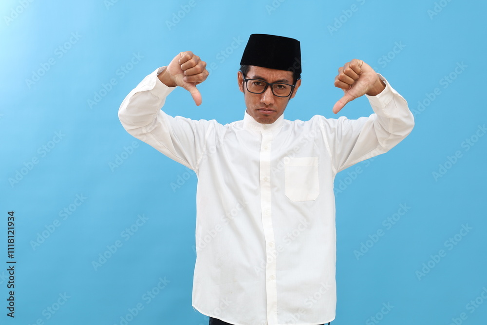 Unhappy Young asian man wearing typical muslim clothes standing while showing thumbs down isolated on blue background Unhappy Young asian man wearing typical muslim clothes standing while showing thumbs down isolated on blue background