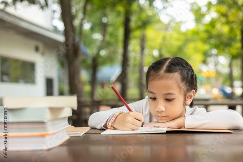 Adorable Asian girl intently writes outdoors, focused on her homework. Surrounded by books, the scene captures childhood learning, concentration, and the joy of studying in outdoor nature.