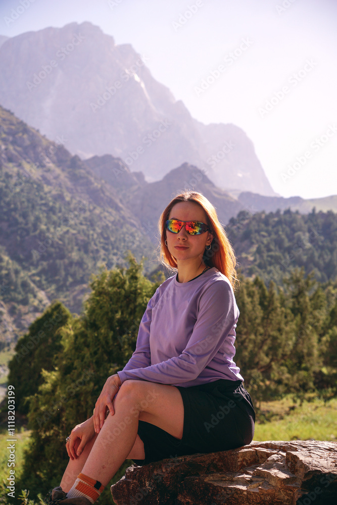 Naklejka premium a young girl with loose red hair and glasses is sitting against the background of mountains. mountain trekking in summer