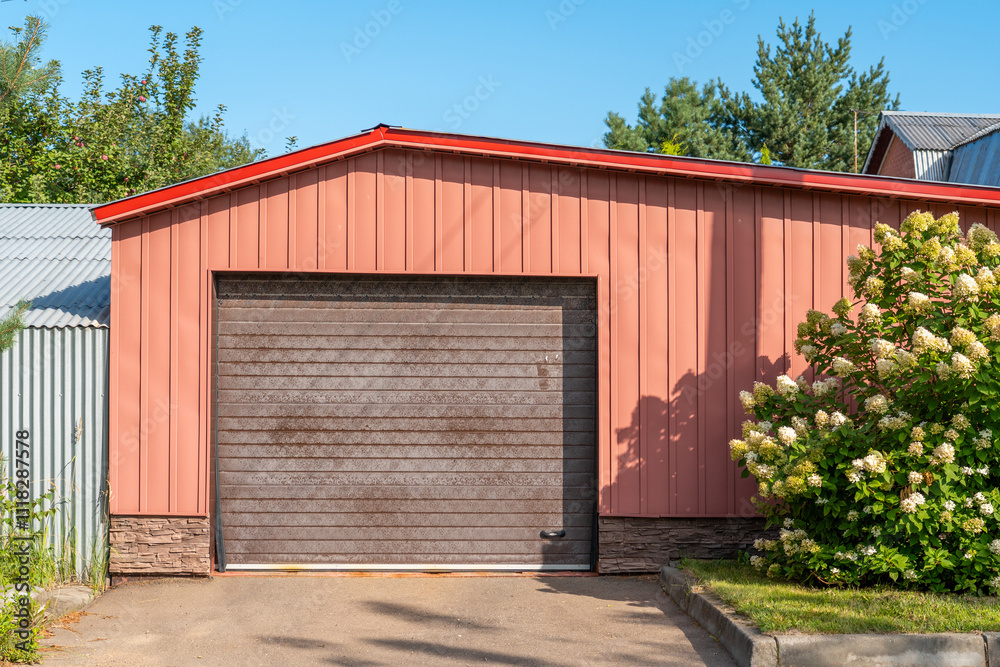Fototapeta premium Bright and inviting garage with a red roof stands against a clear blue sky, bordered by lush green trees and flowering shrubs in full bloom