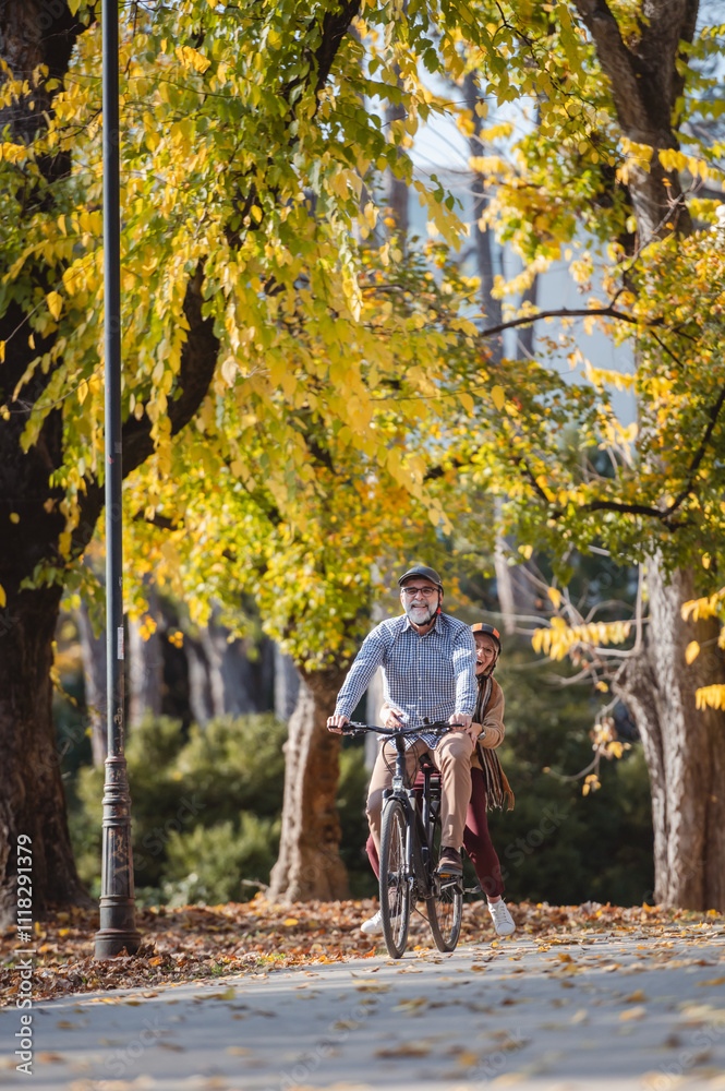 Fototapeta premium Smiling senior couple riding a bike in a sunny autumn park, wearing protective helmets. The man pedals while the woman enjoys the ride sitting on the rear rack