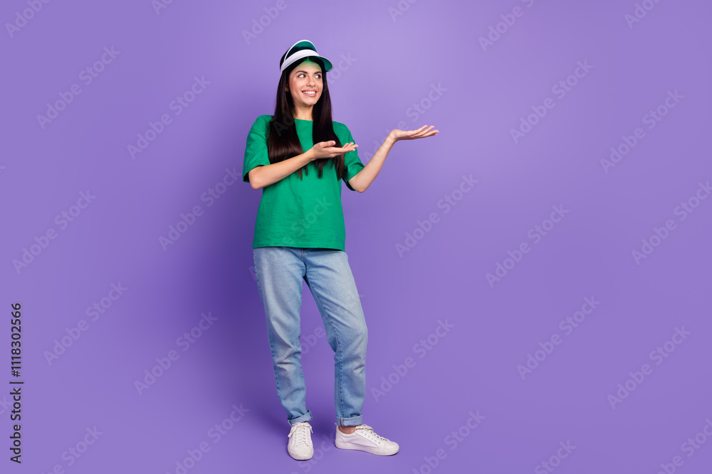 Young woman in casual green t-shirt and jeans posing with open hands on purple background