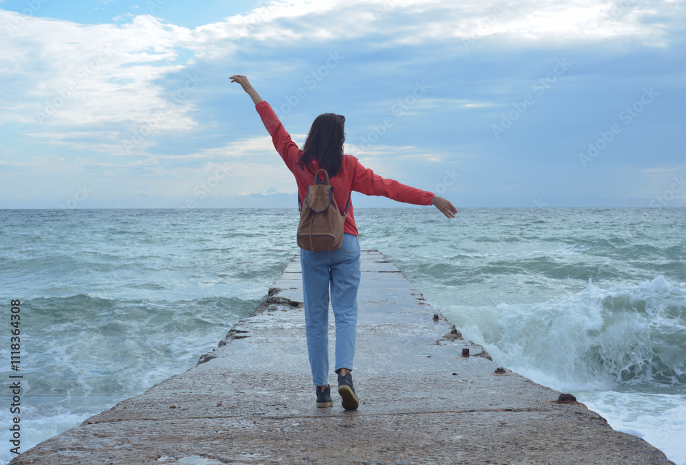 Rear view of a young woman tourist with a backpack standing on the pier and waves hitting the pier in a storm. City of Sudak, Crimea.
