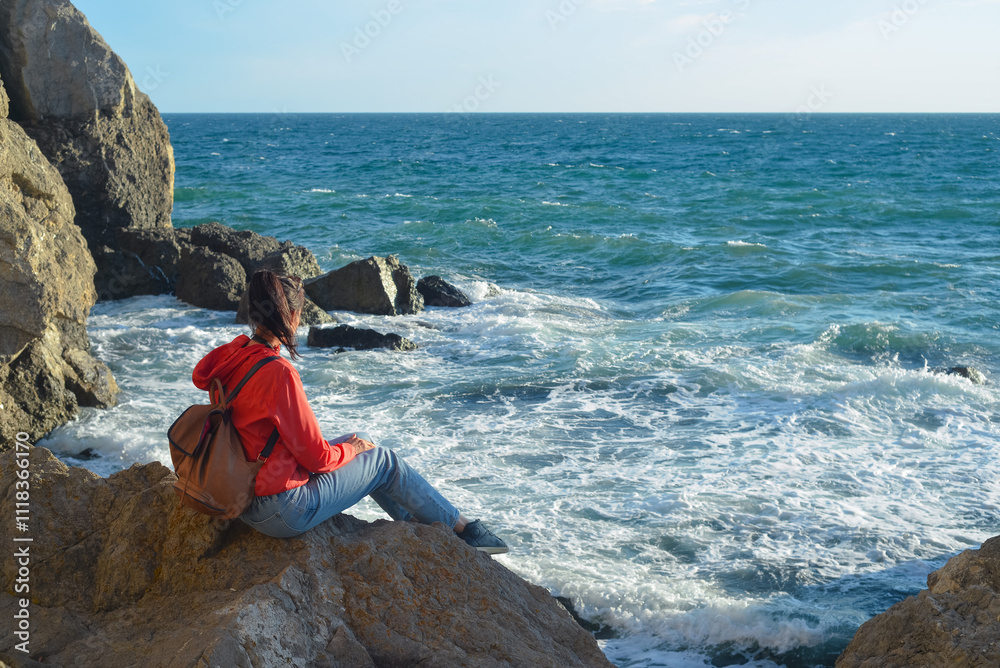 Happy female tourist with a backpack on the top of a mountain, enjoying the sea view, standing on a rock and looking into the distance. The village of Novy Svet in Crimea. Russia