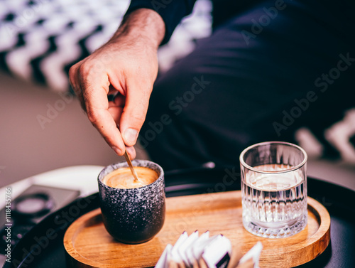 Description: Close-up of a person stirring a cup of espresso on a wooden tray with a glass of water, creating a cozy and stylish coffee moment. Modern café atmosphere.