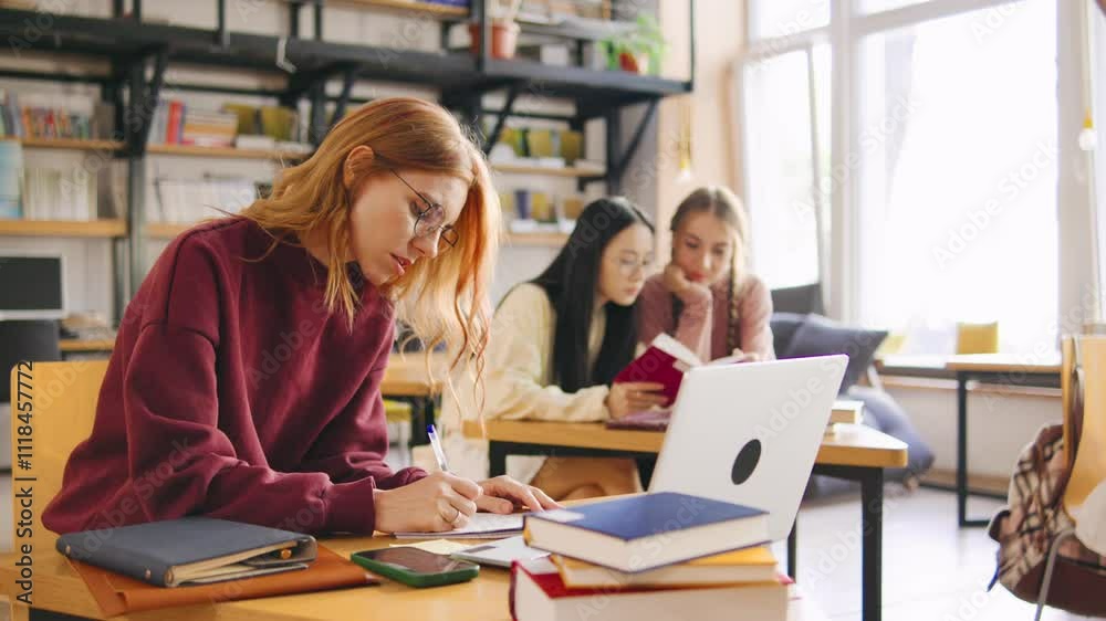 Red-haired female student in glasses sits in the foreground during an online lecture, taking notes in a notebook. Classmates are blurred in the background. Red-haired female student in glasses sits in the foreground during an online lecture, taking notes in a notebook. Classmates are blurred in the background.