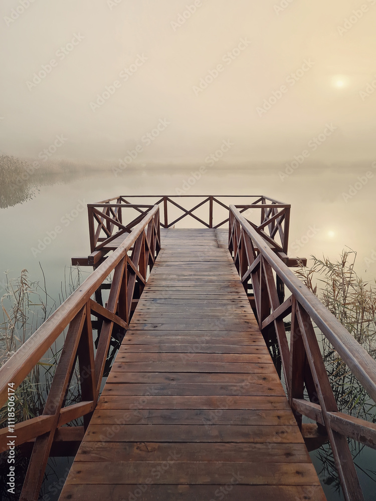 Obraz premium Wooden pier by the lake leading into a foggy scene, surrounded by tall dry reed. The sunrise sun is faintly visible through the mist, creating a serene and tranquil atmosphere. Calm and ethereal scene