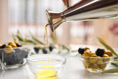 Close-up extra virgin olive oil dripping from a metal cruet into a glass bowl against the background of ripe organic green and black marinated olives on a white table. Mediterranean food concept. photo