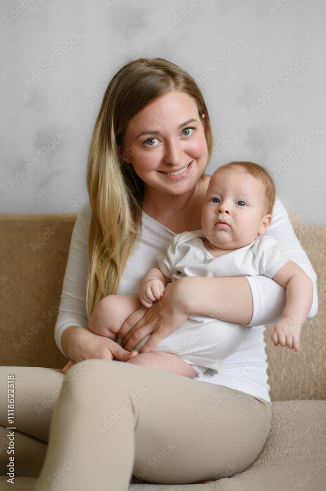 Happy mom sitting on sofa in living room, smiling, holding baby in her arms. Mom and baby in tender moment. Family and motherhood, infancy concept. Vertical Happy mom sitting on sofa in living room, smiling, holding baby in her arms. Mom and baby in tender moment. Family and motherhood, infancy concept. Vertical