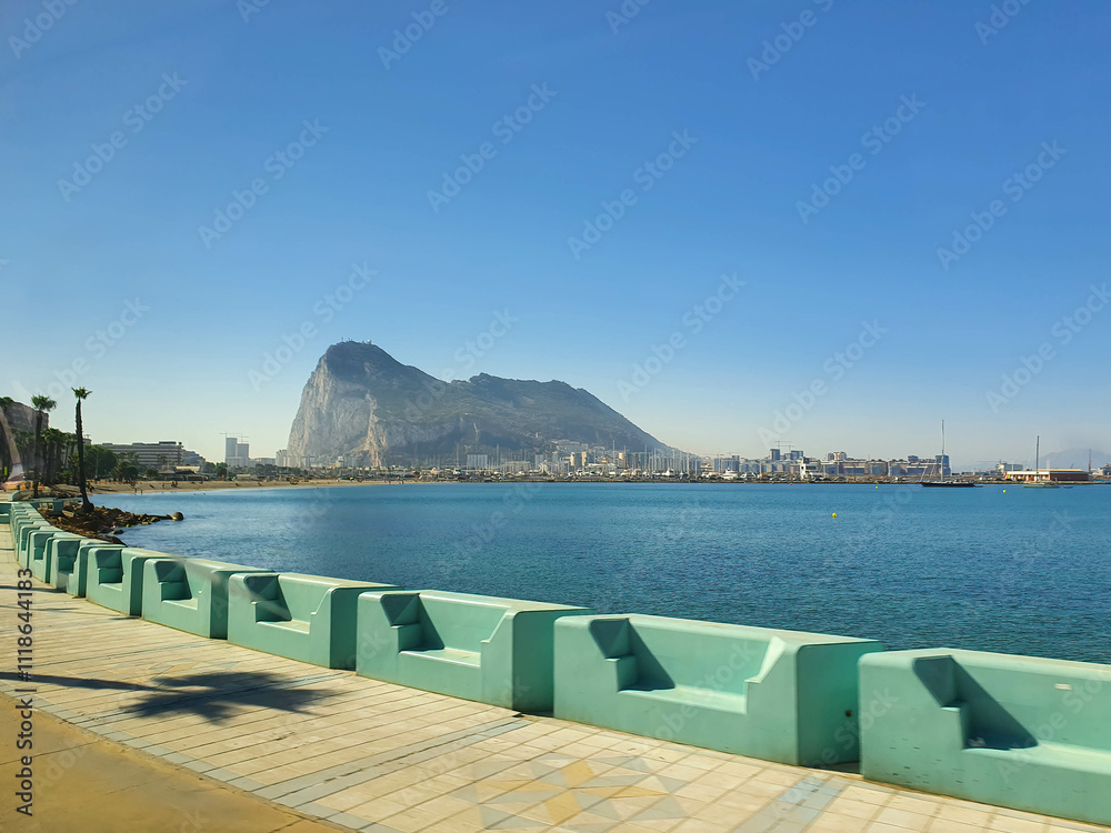 Fototapeta premium Gibraltar rock overseeing the seafront promenade with palm trees and benches on a sunny summer day