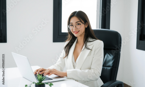 business woman sitting at table with laptop, smiling confidently in modern office setting. Her professional attire and glasses enhance her focused demeanor