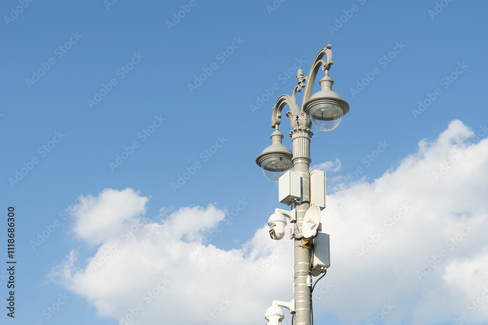 Surveillance cameras with repeater on a lamp post. Carved light pole against a cloudy sky. Surveillance cameras with repeater on a lamp post. Carved light pole against a cloudy sky.
