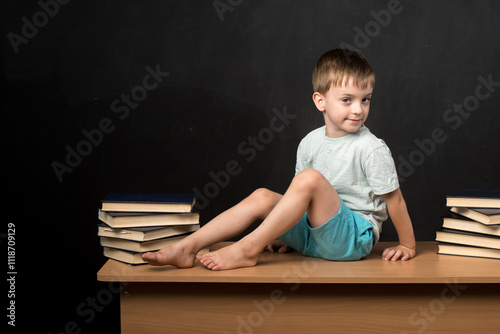 A young boy in casual clothes sits on a wooden desk with stacks of books on both sides, against a blackboard background. Perfect for educational and back-to-school themes photo