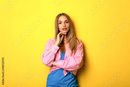 A young woman posing confidently against a vibrant yellow background in a stylish outfit while showcasing her thoughtful expression