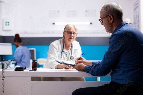 Elderly physician assists retired white man in completing medical forms, both fully focused on his information in hospital. Old doctor seated at clinic desk as pensioner patient writes on clipboard. photo