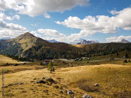 Panorama montano mozzafiato nelle Dolomiti italiane, con colline dorate, una strada tortuosa e un cielo azzurro punteggiato da nuvole bianche. L'immagine cattura la bellezza naturale e la tranquillità photo