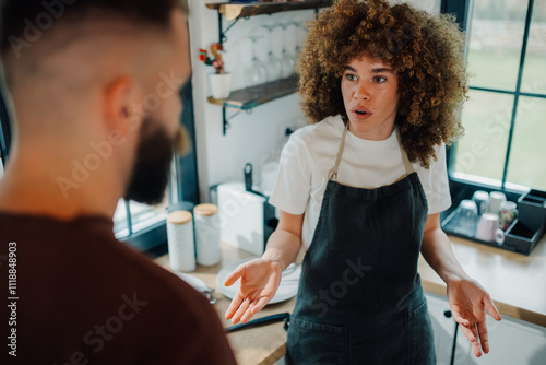 Young frustrated barista woman wearing apron arguing with customer in coffee shop