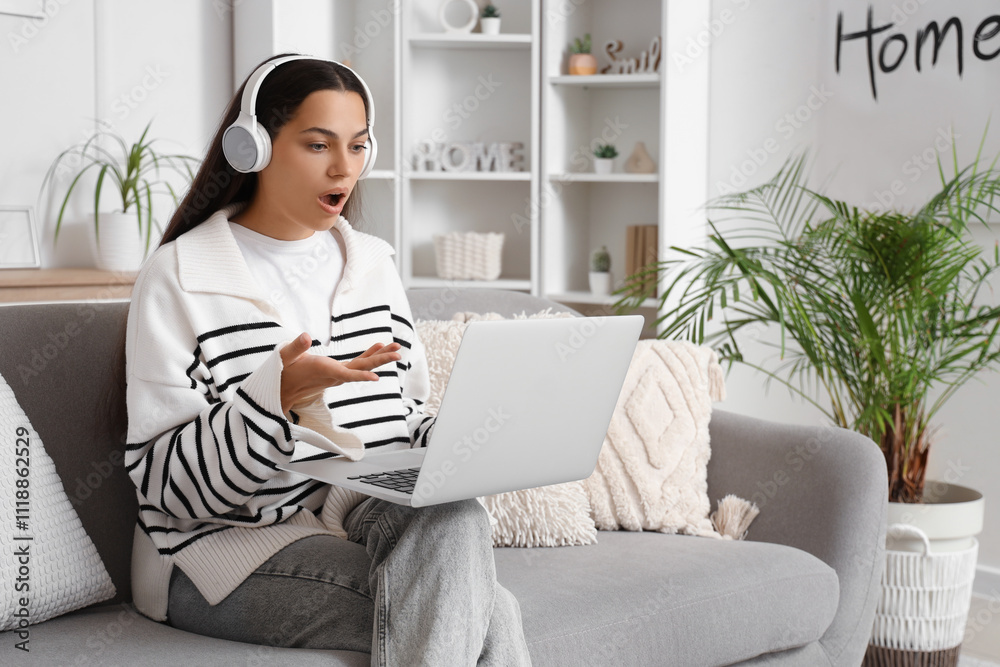 Shocked young woman in headphones with laptop video chatting on sofa at home Shocked young woman in headphones with laptop video chatting on sofa at home