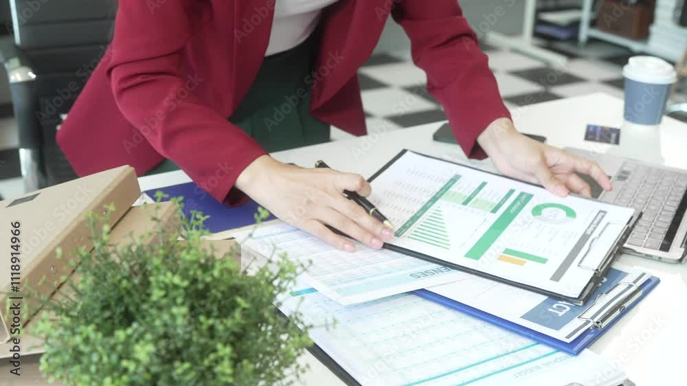 A young Asian woman works diligently at her desk in a bright office room during the day. Surrounded by files, charts, and devices, she strategies on marketing funnels and SEO optimization.