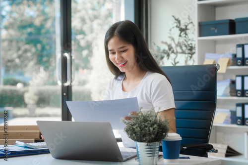 A young Asian woman works at her desk in an office room during the day. Surrounded by financial charts, files, and devices, she consults on credit reports, calculations, and credit risk assessments.