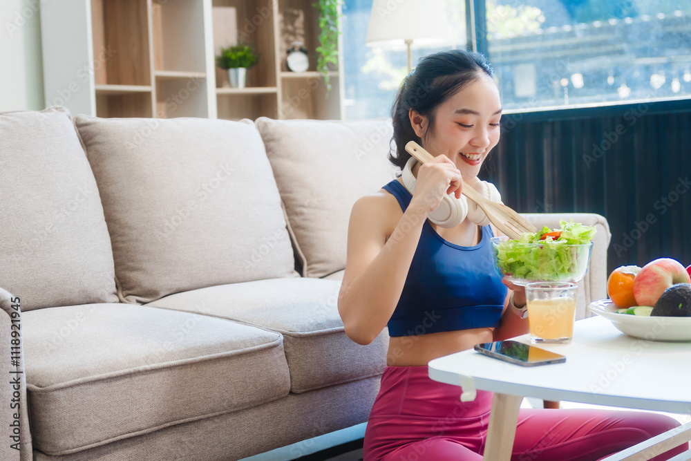 A young Asian housewife enjoys a healthy lifestyle at home, balancing fitness routines with relaxation. She sits on a modern sofa, eating a fresh salad and fruit while embracing healthy habits. A young Asian housewife enjoys a healthy lifestyle at home, balancing fitness routines with relaxation. She sits on a modern sofa, eating a fresh salad and fruit while embracing healthy habits.