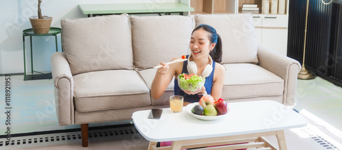 A young Asian housewife enjoys a healthy lifestyle at home, balancing fitness routines with relaxation. She sits on a modern sofa, eating a fresh salad and fruit while embracing healthy habits.