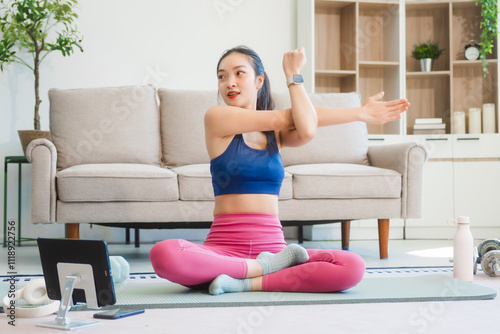 A young Asian housewife sits on sofa in her living room, using fitness app on her mobile with headphones. dumbbells and whey protein, she balances yoga, workouts, vegetarian diet and weight loss.