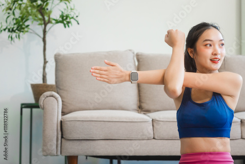 A young Asian housewife sits on sofa in her living room, using fitness app on her mobile with headphones. dumbbells and whey protein, she balances yoga, workouts, vegetarian diet and weight loss.