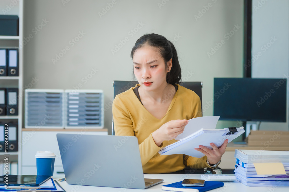 A young Asian businesswoman works alone at a desk with a laptop, stack of papers, and documents. She looks serious and stressed, analyzing financial losses and equity management under daylight. A young Asian businesswoman works alone at a desk with a laptop, stack of papers, and documents. She looks serious and stressed, analyzing financial losses and equity management under daylight.