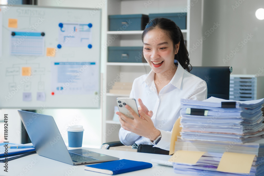 A young Asian businesswoman works alone at a desk with a laptop, stack of papers, and documents. She looks happy and successful, managing accounts and preparing financial statements in daylight. A young Asian businesswoman works alone at a desk with a laptop, stack of papers, and documents. She looks happy and successful, managing accounts and preparing financial statements in daylight.