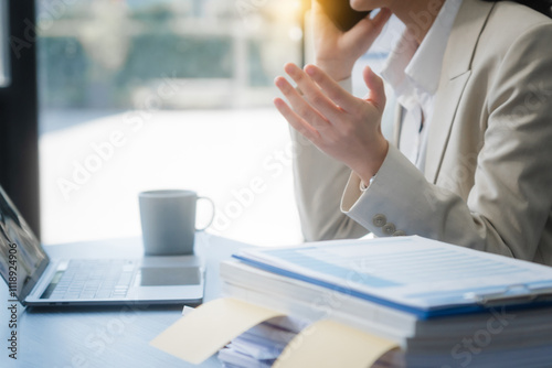 A young Asian businesswoman works alone at a desk with a laptop, stack of papers, and documents. She looks happy and successful, managing accounts and preparing financial statements in daylight. photo