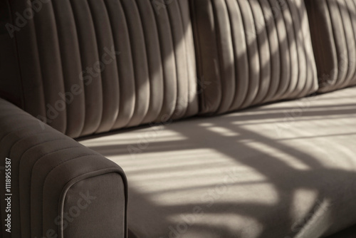 Soft beige velour sofa quilted with straight lines of stitching in shadows and sunlight. Part of the sofa with armrest and pillows close-up with copy space.