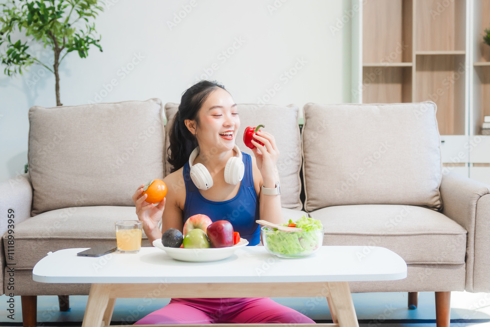 A young Asian housewife enjoys a healthy lifestyle at home, balancing fitness routines with relaxation. She sits on a modern sofa, eating a fresh salad and fruit while embracing healthy habits. A young Asian housewife enjoys a healthy lifestyle at home, balancing fitness routines with relaxation. She sits on a modern sofa, eating a fresh salad and fruit while embracing healthy habits.