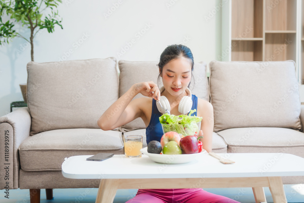 A young Asian housewife enjoys a healthy lifestyle at home, balancing fitness routines with relaxation. She sits on a modern sofa, eating a fresh salad and fruit while embracing healthy habits. A young Asian housewife enjoys a healthy lifestyle at home, balancing fitness routines with relaxation. She sits on a modern sofa, eating a fresh salad and fruit while embracing healthy habits.