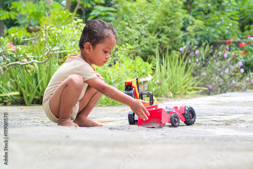 squatting asian toddler holding and playing yellow plastic toy truck on ground