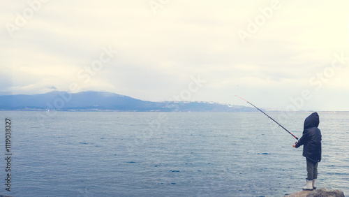 A lone child stands by the lake, fishing under a misty sky.