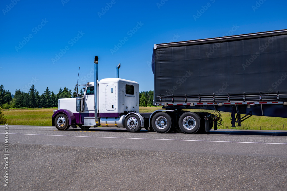 Stylish white and purple classic American big rig semi truck with extended cab transporting cargo in black conestoga flat bad semi trailer running on the summer highway road Stylish white and purple classic American big rig semi truck with extended cab transporting cargo in black conestoga flat bad semi trailer running on the summer highway road