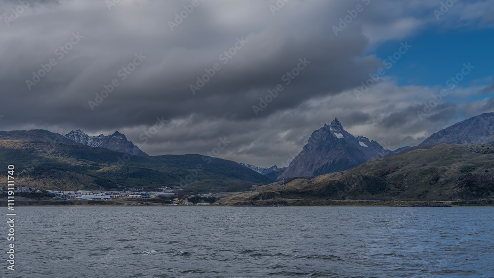 Mountains with snow-capped peaks against the blue sky and clouds. On the ocean shore, at the foot of the green hills, the city of Ushuaia. Ripples on the water. Argentina. Tierra del Fuego Archipelago Mountains with snow-capped peaks against the blue sky and clouds. On the ocean shore, at the foot of the green hills, the city of Ushuaia. Ripples on the water. Argentina. Tierra del Fuego Archipelago