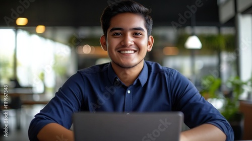 Young man in blue shirt sitting at desk with laptop, smiling and looking at camera, working remotely in office setting