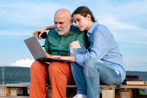 Old grandad and young grandchild using laptop. Young girl and old father using laptop, surfing internet outdoor. Younger generation. Caring about older relatives. Old young with laptop concept.